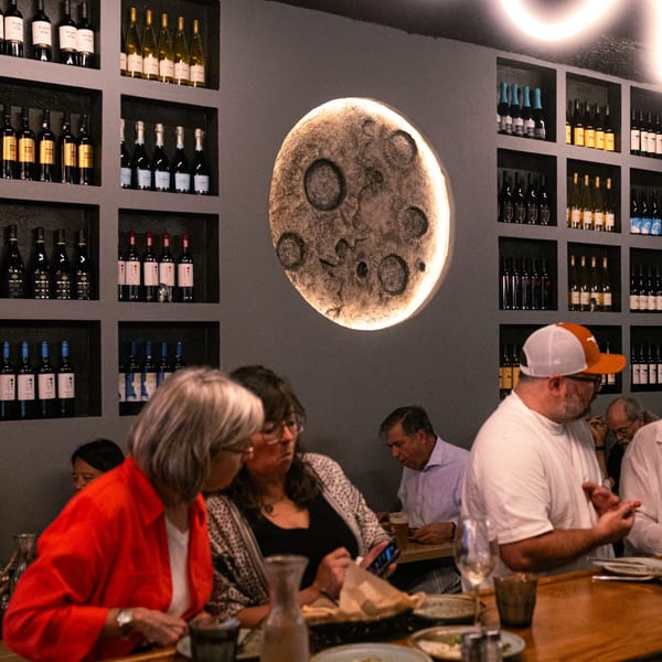 Diners seated at tables in The Parthenon restaurant, with a moon-like wall decoration and wine bottles.