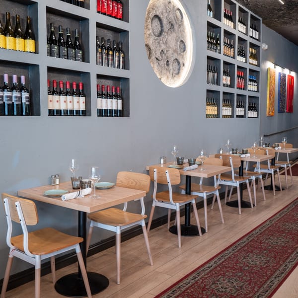 The Parthenon restaurant interior with set tables, a moon art piece, and wine bottles displayed on shelves.