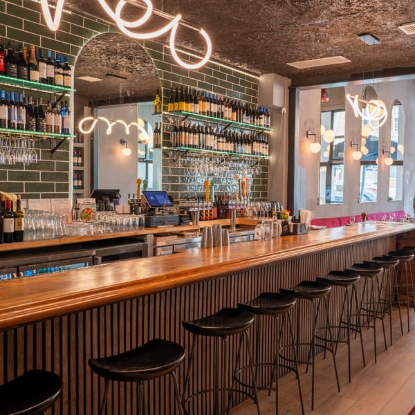 A long, well-stocked bar with bottles, glasses, beer taps, and black bar stools at The Parthenon.