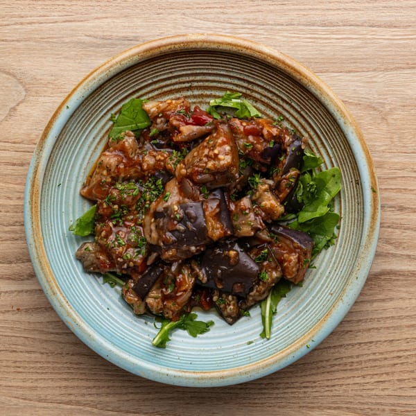 A blue and brown bowl filled with Greek eggplant stew, garnished with fresh parsley and greens.