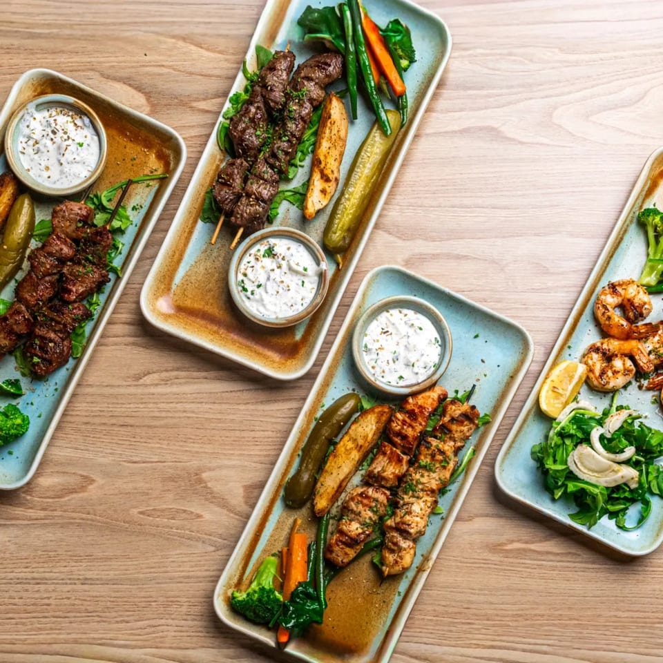 Four Greek platters with various grilled meats, shrimp, vegetables, and tzatziki sauce on a wooden table.