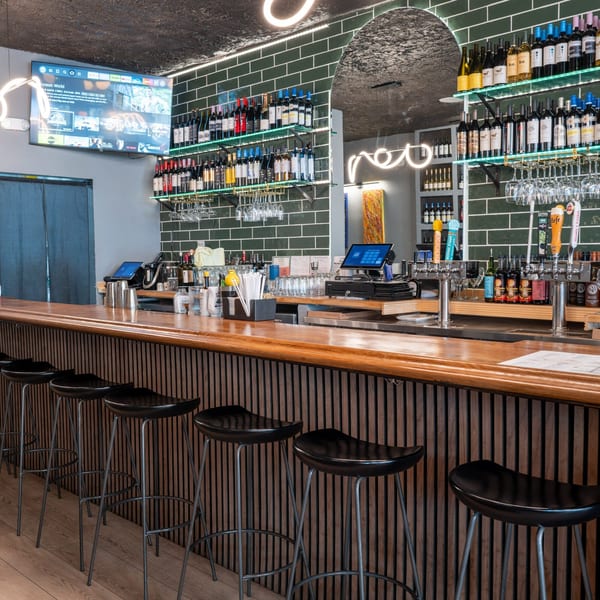 A modern bar area with a wooden counter, black stools, green tiled walls, and shelves of wine bottles.