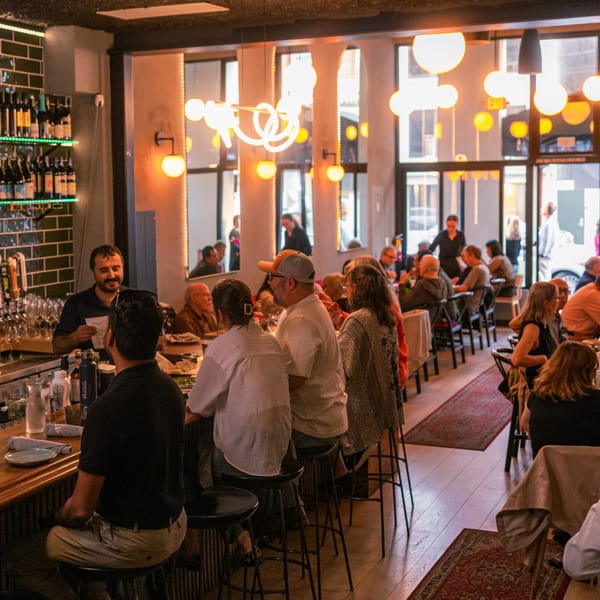 A lively Greek restaurant interior with patrons dining at tables and a bar, illuminated by warm lighting.